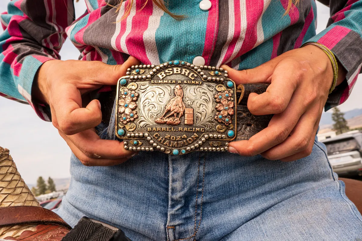 Hands holding a silver and turquoise barrel racing belt buckle over blue jeans.