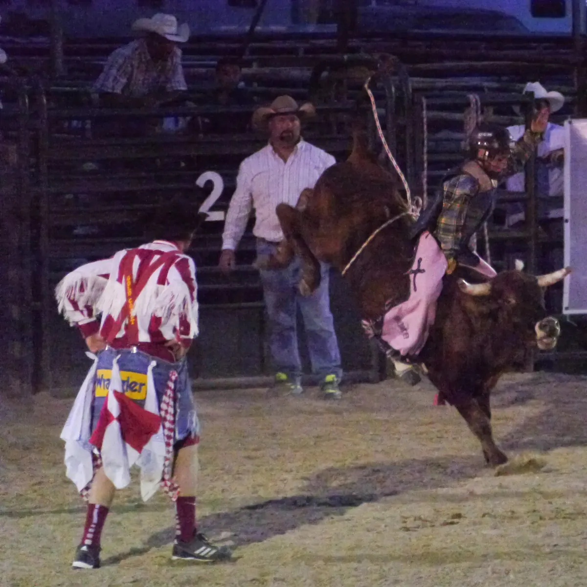 A rider in a helmet and chaps on a bucking brown bull at a night rodeo in Texas, with a rodeo clown watching.